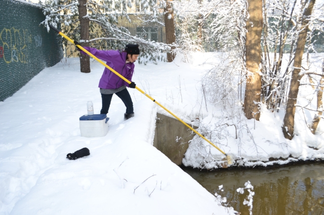 A woman uses a long pole reaching from a bridge into the water on a snowy day.
